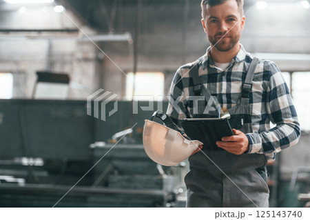 Black colored tablet in hands. Factory male worker in uniform is indoors Black colored tablet in hands. Factory male worker in uniform is indoors 125143740