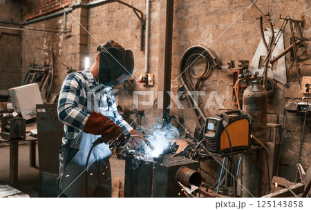 In protective mask. Welding the iron. Factory male worker in uniform is indoors In protective mask. Welding the iron. Factory male worker in uniform is indoors 125143858
