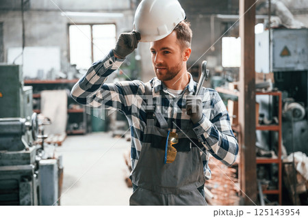 Standing in the warehouse with artificial lighting. Factory male worker in uniform is indoors Standing in the warehouse with artificial lighting. Factory male worker in uniform is indoors 125143954