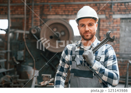 Standing in grey uniform and in white hard hat. Factory male worker is indoors 125143986