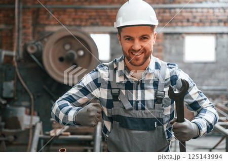 Standing and showing wrench. Factory male worker in uniform is indoors 125143994