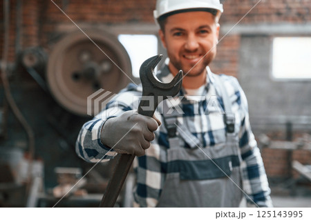 Standing and showing wrench. Factory male worker in uniform is indoors 125143995
