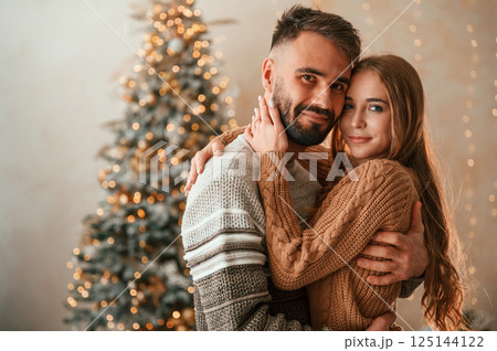 Standing against the fir with garlands on it. Lovely young couple are celebrating New Year at home Standing against the fir with garlands on it. Lovely young couple are celebrating New Year at home 125144122