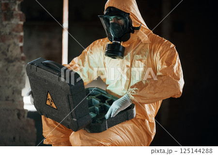 Case with blue glowing light. Hazard materials. Man dressed in chemical protection suit in the ruins of the post apocalyptic building Case with blue glowing light. Hazard materials. Man dressed in chemical protection suit in the ruins of the post apocalyptic building 125144288