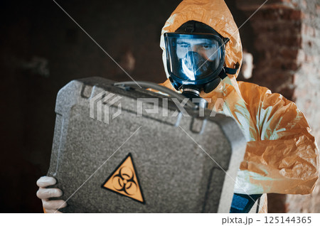 Blue light is going from hard case that is with hazard sign. Man dressed in chemical protection suit in the ruins of the post apocalyptic building Blue light is going from hard case that is with hazard sign. Man dressed in chemical protection suit in the ruins of the post apocalyptic building 125144365