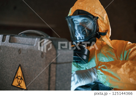 Blue light is going from hard case that is with hazard sign. Man dressed in chemical protection suit in the ruins of the post apocalyptic building Blue light is going from hard case that is with hazard sign. Man dressed in chemical protection suit in the ruins of the post apocalyptic building 125144366