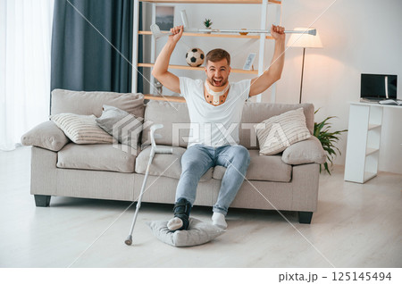 Holding crutch hands while sitting on sofa and watching sport match. Man is at home indoors. Having injuries 125145494