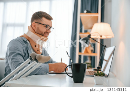 By the table with laptop. Man with crutches is at home indoors. Having injuries 125145511