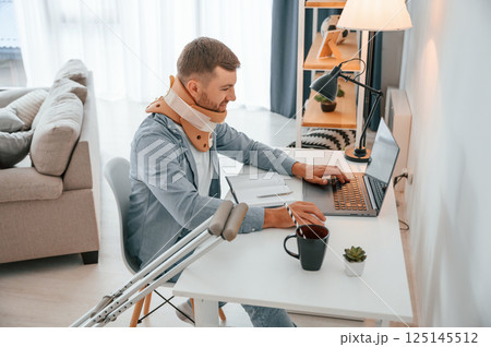 By the table with laptop. Man with crutches is at home indoors. Having injuries 125145512