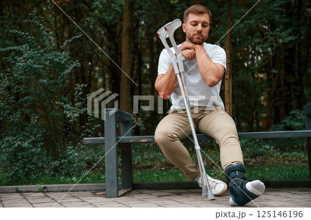 Sitting, feeling bored and sad. Man with crutches is in the park outdoors. Having leg injury 125146196