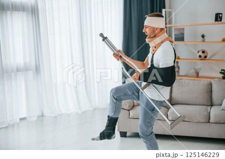 Pretending of playing guitar on crutches. Man is at home indoors. Having injuries Pretending of playing guitar on crutches. Man is at home indoors. Having injuries 125146229