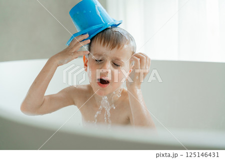Young child having fun during bath time, playfully pouring water over their head using a blue toy bucket. Facial expression shows joy and excitement in a bright bathroom.  125146431