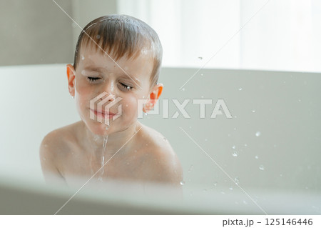 Young boy with eyes closed, feeling calm under a stream of water in a bathtub. Water droplets are visible around him, creating a sense of playful serenity.  125146446