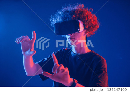 In virtual reality glasses. Young man with curly hair is indoors illuminated by neon lighting 125147100