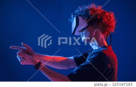 Playing shooter game in VR glasses. Young man with curly hair is indoors illuminated by neon lighting 125147103