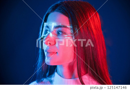 In futuristic eyewear. Young beautiful woman is indoors in the studio illuminated by neon light 125147146