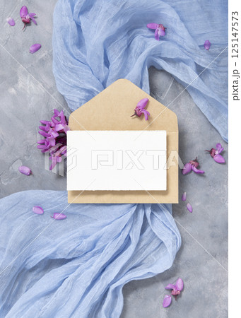 Card and envelope on fabric near wild purple flowers top view on gray, spring wedding mockup 125147573