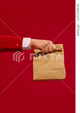 Hand holding kraft paper bag against bold red background. Express delivery with eco-friendly packaging. 125149016