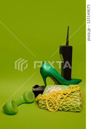 High heel, rotary phone and brick mobile phone arranged on green backdrop with tangled yellow mesh. 125149075