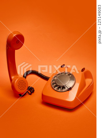 Orange vintage rotary dial telephone with receiver slightly lifted off its base is positioned against monochromatic orange backdrop. 125149083