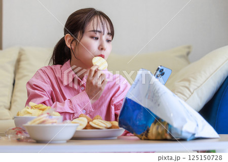 スマホを見ながらお菓子を食べる女性 スマホを見ながらお菓子を食べる女性 125150728