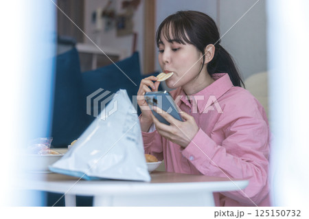 スマホを見ながらお菓子を食べる女性 スマホを見ながらお菓子を食べる女性 125150732
