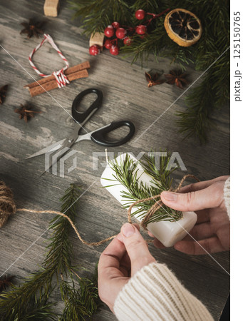 Female hands making atmospheric Christmas decoration with candle, spruce needles, jute twine on a wooden table, vertical image. 125150765
