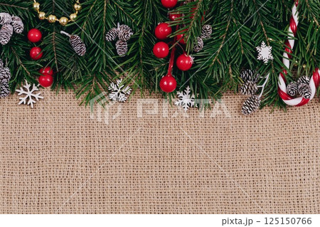 Christmas decorative upper border with green spruce, cones, candy cane and holly berries on canvas background, top view, copy space. 125150766