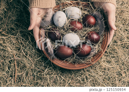 Female hands with clay plate of Easter painted eggs among hay background, close-up, copy space. Female hands with clay plate of Easter painted eggs among hay background, close-up, copy space. 125150934