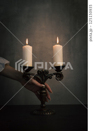 Female hand in white vintage dress sleeve take old brass candelabrum with burning candles from table in the dark, vertical image. 125150935