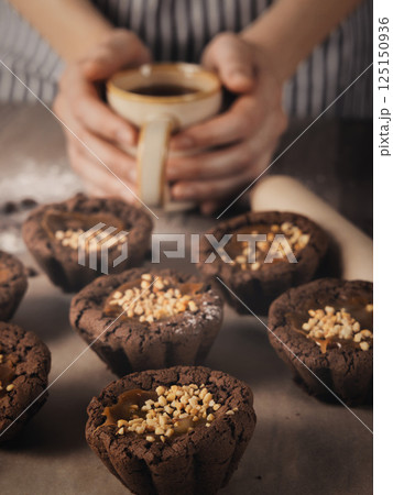 Female hands hold coffee mug and freshly baked muffins on baking paper on a table, vertical image. Female hands hold coffee mug and freshly baked muffins on baking paper on a table, vertical image. 125150936