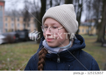 Teenager Girl Walking Alone on City Street. Portrait of 14 years old girl wearing eyeglasses, wool hat and winter jacket walks around the city. Urban Style. Rainy mood weather. Cold season. One person Teenager Girl Walking Alone on City Street. Portrait of 14 years old girl wearing eyeglasses, wool hat and winter jacket walks around the city. Urban Style. Rainy mood weather. Cold season. One person 125152657