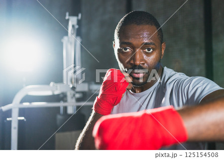 Workout in gym. African man fighter ready for fight punching with boxing wraps protective bandages to camera. Strong man training punches looking concentrated straight preparing for boxing sparring Workout in gym. African man fighter ready for fight punching with boxing wraps protective bandages to camera. Strong man training punches looking concentrated straight preparing for boxing sparring 125154508