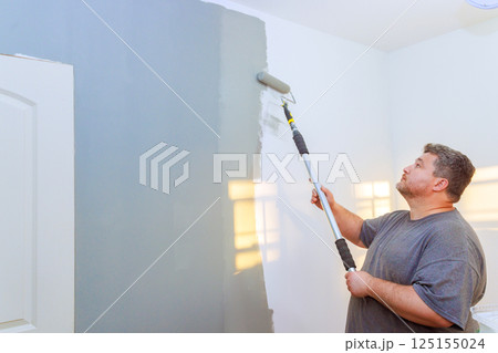Worker is carefully applying gray paint to wall with roller while standing in well lit room. Worker is carefully applying gray paint to wall with roller while standing in well lit room. 125155024