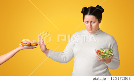 Say No To Obesity. Annoyed Plump Woman Showing Stop Gesture To Offered Plate With Donuts And Eating Fresh Vegetable Salad, Yellow Background Say No To Obesity. Annoyed Plump Woman Showing Stop Gesture To Offered Plate With Donuts And Eating Fresh Vegetable Salad, Yellow Background 125172868
