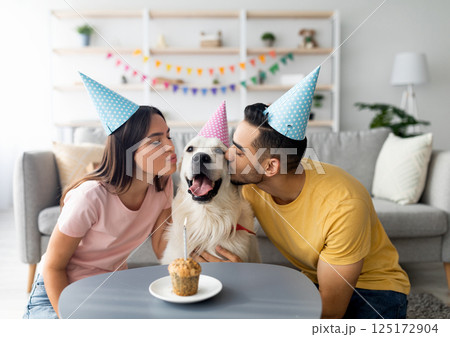 Young interracial couple in party hats celebrating their dog's birthday, kissing their pet at home. Millennial owners presenting golden retriever with small b-day cake indoors 125172904
