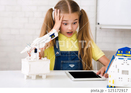 A young girl with pigtails looks surprised and concerned as she works on a robotics project. She is surrounded by robot parts and a tablet, suggesting she is following instructions A young girl with pigtails looks surprised and concerned as she works on a robotics project. She is surrounded by robot parts and a tablet, suggesting she is following instructions 125172917