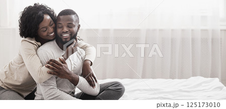 Loving wife cuddling husband from the back while sitting on bed together, enjoying weekend. Copy space 125173010