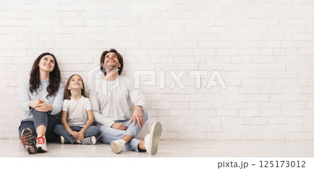 Happy young family of three sitting on floor and looking upwards, dreaming about something, leaning on white brick wall, free space 125173012