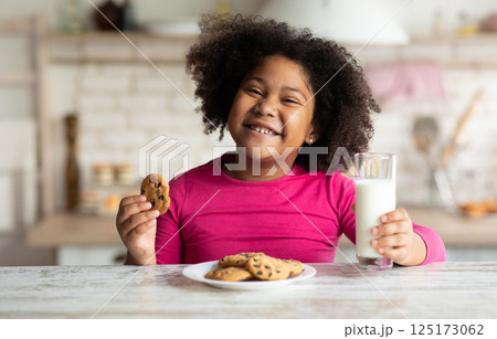 Portrait Of Cute African American Girl Enjoying Cookies And Milk In Kitchen, Pretty Black Child Eating Snack At Home, Preschool Kid Having A Bite With Fresh Baked Biscuits And Calcium Drink, Closeup 125173062