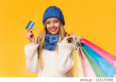 Smiling young winter woman in blue hat recommending credit card, posing with shopping bags and looking at camera, pink background, free space 125173066