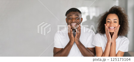 A cheerful African American couple stands side by side in a bright room, smiling widely while playfully touching their faces, radiating happiness and a carefree atmosphere during the day. 125173104