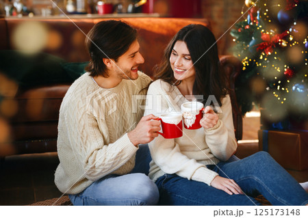 Cheerful man and woman having xmas cocoa by Christmas tree at home 125173148