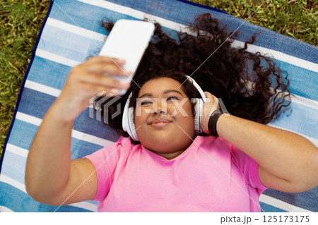A young woman lies on a blue striped mat, wearing headphones and smiling as she takes a selfie. It a sunny day, and she enjoys her music in a grassy outdoor area. 125173175