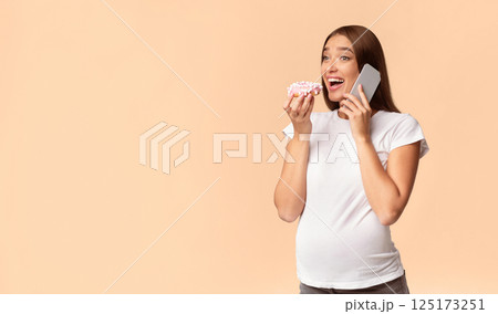 Excited Pregnant Woman Chatting On Cellphone Eating Doughnut Standing On White Studio Background. Copy Space 125173251