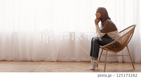 Morning Harmony. Muslim girl in hijab relaxing in wicker chair at home, resting head on hands and daydreaming against window, side view 125173262