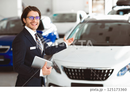 Car Sales. Smiling Salesman In Glasses Showing New Auto Standing In Automobile Dealership Showroom. Selective Focus Car Sales. Smiling Salesman In Glasses Showing New Auto Standing In Automobile Dealership Showroom. Selective Focus 125173360