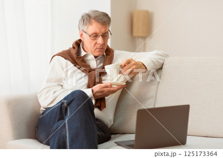 Senior Man Using Laptop Computer Having Coffee Sitting On Sofa At Home On Weekend. Selective Focus 125173364