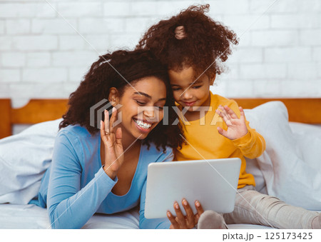 Black Mother And Daughter Using Digital Tablet Making Video Call And Waving Hello Lying In Bed At Home. Selective Focus Black Mother And Daughter Using Digital Tablet Making Video Call And Waving Hello Lying In Bed At Home. Selective Focus 125173425