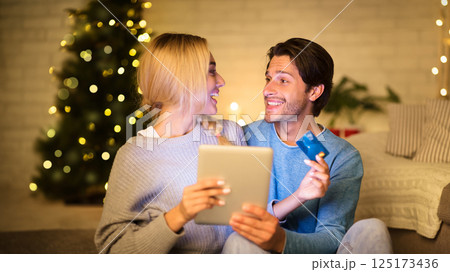 Couple buying Christmas gifts online, using tablet and credit card, sitting in front of Christmas tree 125173436